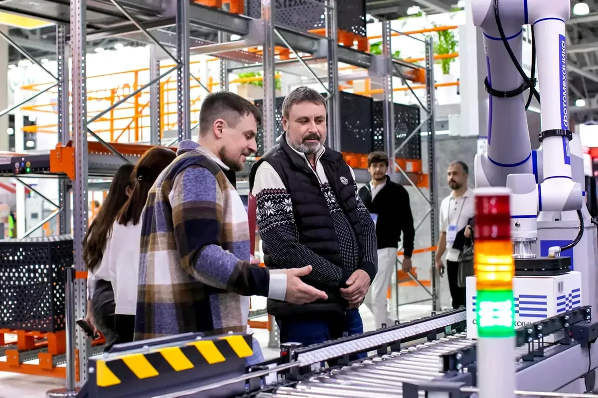 People beside a roller conveyor and robot arm in a warehouse display area.